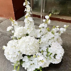 May include: A large, dense floral arrangement featuring clusters of white hydrangea blossoms and tall, slender stalks of white delphinium flowers. The bouquet includes small green stems and leaves interspersed throughout the white petals.