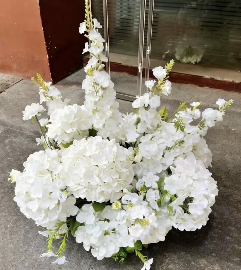 May include: A large, dense floral arrangement featuring clusters of white hydrangea blossoms and tall, slender stalks of white delphinium flowers. The bouquet includes small green stems and leaves interspersed throughout the white petals.