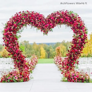 May include: A heart-shaped floral arch, approximately 8 feet wide and 8 feet high, made of red and pink flowers and greenery. The archway is set up outdoors for a wedding or event. The text "ArchFlowers W8xH8 ft" is visible.