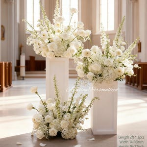 May include: Three white floral arrangements on white pedestals. The arrangements feature roses, hydrangeas, and other white flowers. The pedestals are rectangular and placed in a church setting. The text "Angelina Personalized" is visible.
