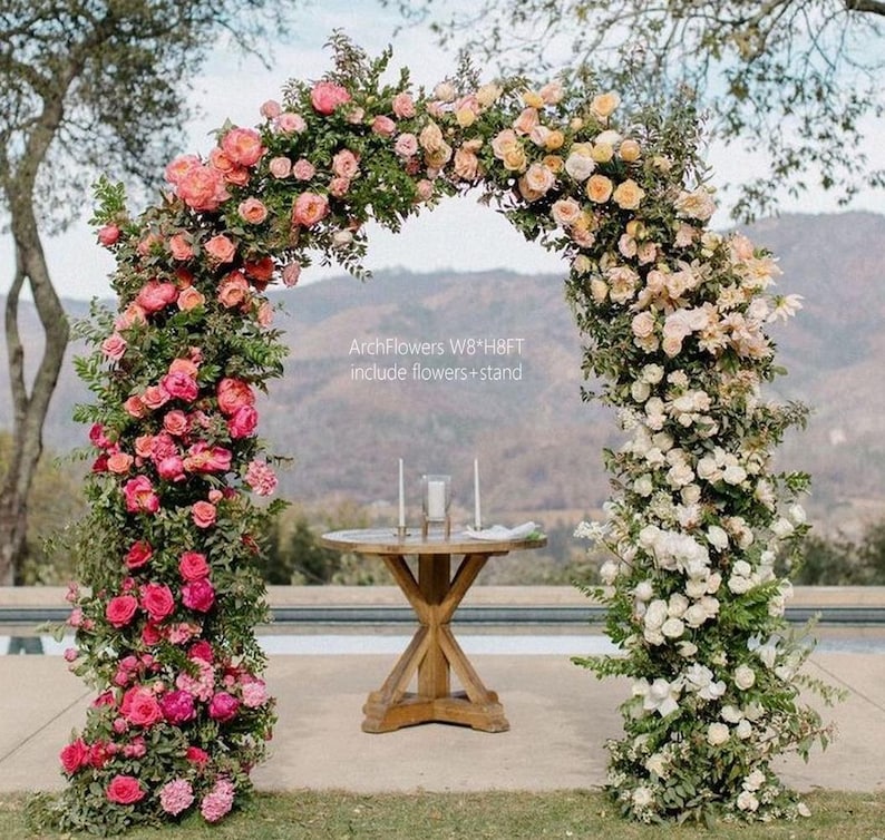 K&ouml;nnte beinhalten: Ein dekorativer Blumenbogen, geschm&uuml;ckt mit rosa, pfirsichfarbenen und wei&szlig;en Rosen und Gr&uuml;n. Der Bogen ist etwa 244 cm breit und 244 cm hoch. Vorne steht ein Holztisch mit Kerzen. Der Text auf dem Bild lautet "ArchFlowers W8*H8FT include flowers + stand."