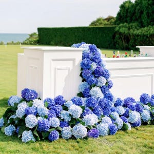 May include: A white outdoor bar decorated with cascading blue and purple hydrangeas. The bar is set on a green lawn with a hedge and ocean in the background. Bottles are visible on the bar top.