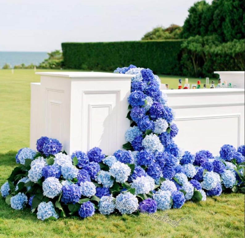 May include: A white outdoor bar decorated with cascading blue and purple hydrangeas. The bar is set on a green lawn with a hedge and ocean in the background. Bottles are visible on the bar top.