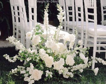 以下が含まれることがあります： 白いバラやその他の白い花と緑の葉のフラワーアレンジメントが芝生の上にあります。アレンジメントの長さは61cm、幅は36〜41cm、高さは41〜61cmです。背景には白い椅子があります。