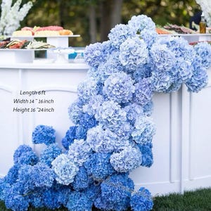 May include: A display of blue hydrangea flowers cascading over a white surface. The arrangement has a length of 6 ft, a width of 14-16 inches, and a height of 16-24 inches. The flowers are a mix of light and dark blue hues, creating a visually appealing floral arrangement.
