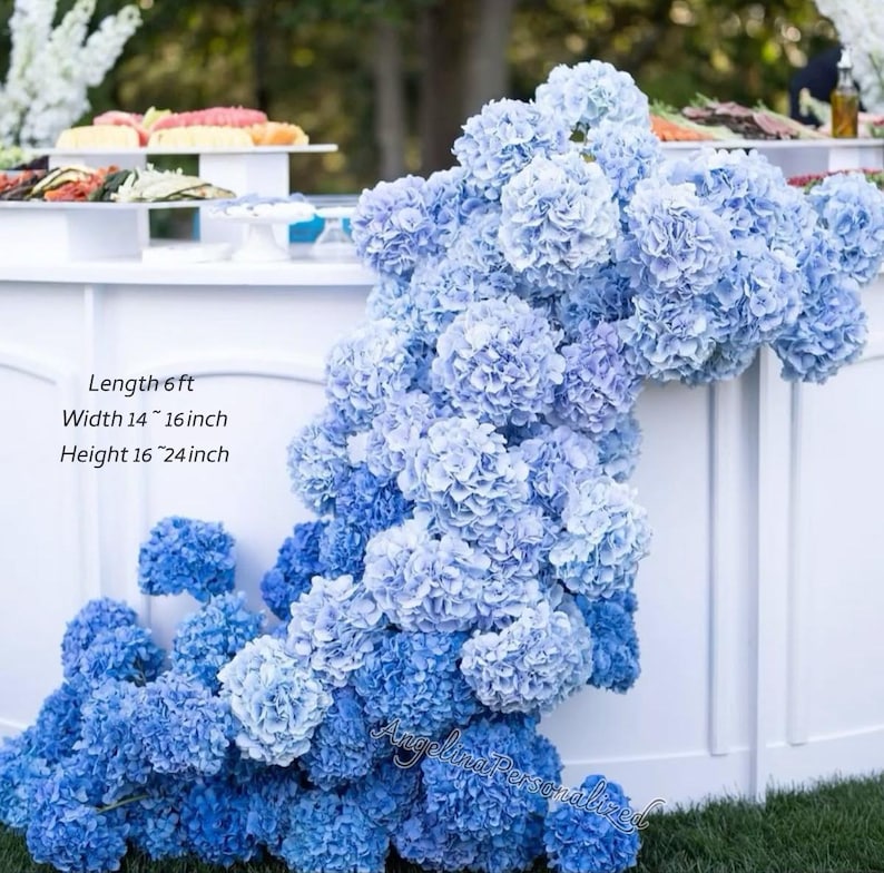 May include: A display of blue hydrangea flowers cascading over a white surface. The arrangement has a length of 6 ft, a width of 14-16 inches, and a height of 16-24 inches. The flowers are a mix of light and dark blue hues, creating a visually appealing floral arrangement.