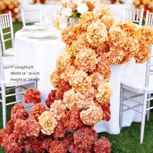 May include: A floral arrangement of orange and red hydrangeas cascading over a white tablecloth. The arrangement is approximately 6 feet long, 14 to 16 inches wide, and 16 to 24 inches high. The flowers are arranged on a table with white chairs.