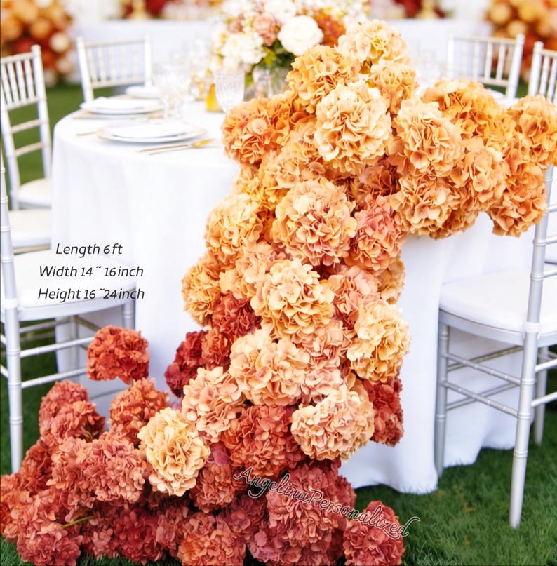 May include: A floral arrangement of orange and red hydrangeas cascading over a white tablecloth. The arrangement is approximately 6 feet long, 14 to 16 inches wide, and 16 to 24 inches high. The flowers are arranged on a table with white chairs.