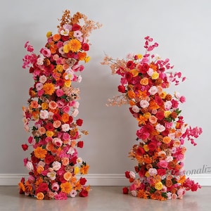 Könnte beinhalten: Zwei Blumenarrangements in einem Raum. Jedes Arrangement ist eine vertikale Säule aus bunten Blumen, darunter Rosen und Dahlien, in Rot-, Orange-, Rosa- und Gelbtönen. Die Blumen sind in einer gebogenen Form angeordnet.