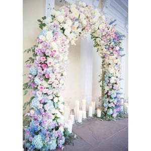 May include: A wedding arch decorated with a gradient of pastel flowers, starting with white and transitioning to light blue. The arch is made of greenery and flowers, including roses, hydrangeas, and other blooms. The arch is set up in front of a white wall with candles on the ground.