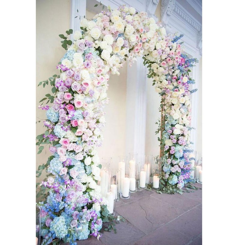 May include: A wedding arch decorated with a gradient of pastel flowers, starting with white and transitioning to light blue. The arch is made of greenery and flowers, including roses, hydrangeas, and other blooms. The arch is set up in front of a white wall with candles on the ground.