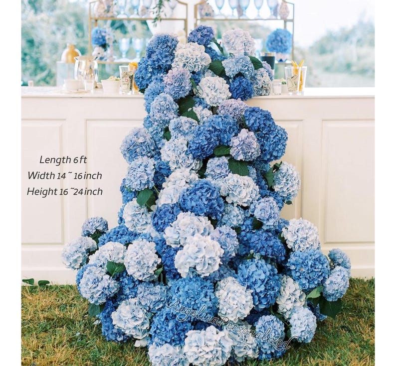 May include: A display of blue and white hydrangeas, with the text "Length 6 ft, Width 14-16 inch, Height 16-24 inch". The flowers are arranged in a cascading design, creating a lush and vibrant floral arrangement. The flowers are on a white surface.