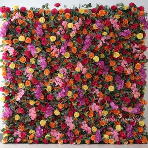 May include: A floral wall backdrop with a variety of artificial flowers in shades of pink, orange, yellow, and red. The flowers are arranged in a dense, lush pattern, creating a vibrant and colorful display.