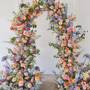 May include: A floral archway arrangement featuring a variety of colorful flowers in shades of pink, orange, yellow, purple, and blue. The arch is composed of roses, hydrangeas, and other blooms, creating a vibrant and elegant display, perfect for weddings or special events.