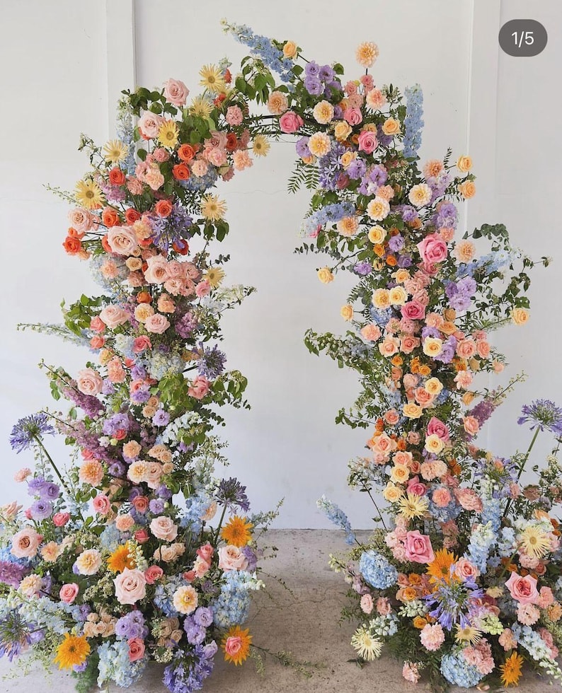 May include: A floral archway arrangement featuring a variety of colorful flowers in shades of pink, orange, yellow, purple, and blue. The arch is composed of roses, hydrangeas, and other blooms, creating a vibrant and elegant display, perfect for weddings or special events.