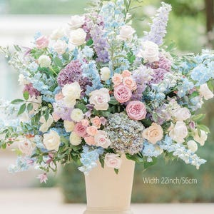 Puede incluir: Un gran arreglo floral en un jarrón crema. El ramo presenta flores blancas, rosas, azules y moradas. El arreglo mide 56 cm de ancho. Ideal para bodas o decoración del hogar.