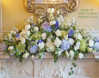 Puede incluir: Arreglo floral con rosas blancas, hortensias azules y otras flores. El arreglo se exhibe sobre una repisa blanca. El texto indica una longitud de 122 cm, un ancho de 36-41 cm y una altura de 41-56 cm.