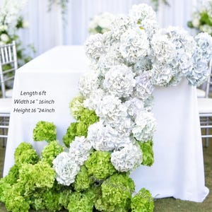 May include: A white tablecloth is adorned with cascading white and light blue hydrangea blooms, accented by vibrant green hydrangeas at the base. The table is 6 ft long, 14-16 inches wide, and 16-24 inches high. The scene suggests a wedding or event setting.