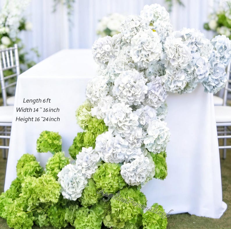 May include: A white tablecloth is adorned with cascading white and light blue hydrangea blooms, accented by vibrant green hydrangeas at the base. The table is 6 ft long, 14-16 inches wide, and 16-24 inches high. The scene suggests a wedding or event setting.