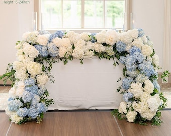 Puede incluir: Un arco floral compuesto por hortensias blancas y azules y vegetación. El arco mide aproximadamente 3 metros de largo, 36-41 cm de ancho y 41-61 cm de alto. Las flores están dispuestas sobre una superficie blanca, con una ventana al fondo.