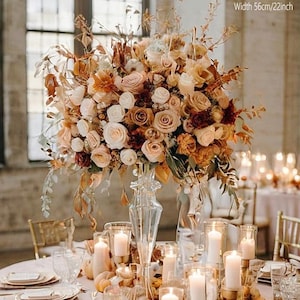Op de afbeelding: Een groot bloemstuk met rozen en andere bloemen in crème, perzik en bruin, gepresenteerd in een heldere glazen vaas. De opstelling is 56 cm breed. De tafelsetting omvat kaarsen, borden en glaswerk, wat een elegante sfeer creëert.