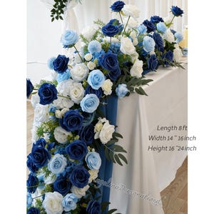 May include: A floral table runner featuring white, light blue, and navy blue roses and other flowers. The arrangement is on a white tablecloth. The text on the image says: Length 8 ft, Width 14-16 inch, Height 16-24 inch.