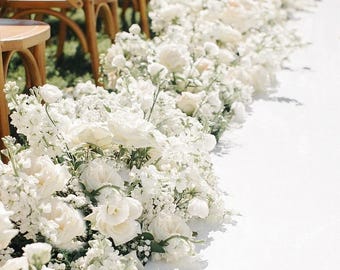Op de afbeelding: Een pad omzoomd met weelderige, witte bloemen, waaronder rozen en andere bloesems, waardoor een bloemenrand ontstaat. Houten stoelen zijn zichtbaar op de achtergrond. De tekst geeft aan dat de bloemstuk 61 cm lang, 36-41 cm breed en 41-56 cm hoog is.