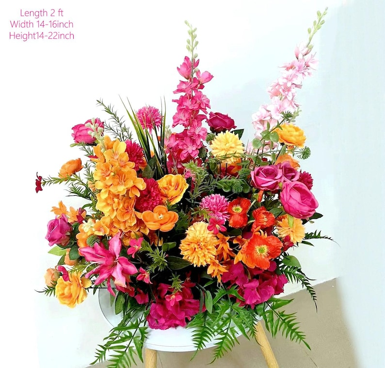 May include: A vibrant floral arrangement featuring a variety of colorful flowers in shades of pink, orange, and yellow. The arrangement is approximately 2 feet long, 14-16 inches wide, and 14-22 inches high. The flowers are arranged in a white container.