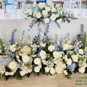 Puede incluir: Un tel&oacute;n de fondo para una ceremonia de boda con flores blancas y azules. Las flores est&aacute;n dispuestas en un dise&ntilde;o en cascada, con rosas blancas, hortensias azules y lisianthus blancos. El tel&oacute;n de fondo tiene 1,52 metros de largo, 35,56 a 40,64 cent&iacute;metros de ancho y 40,64 a 55,88 cent&iacute;metros de alto.