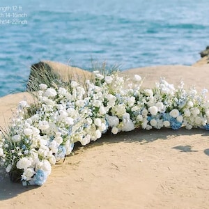 May include: A floral arrangement featuring white and blue flowers, with a length of 12 ft, a width of 14-16 inches, and a height of 14-22 inches. The flowers are arranged on a sandy surface with the ocean in the background.