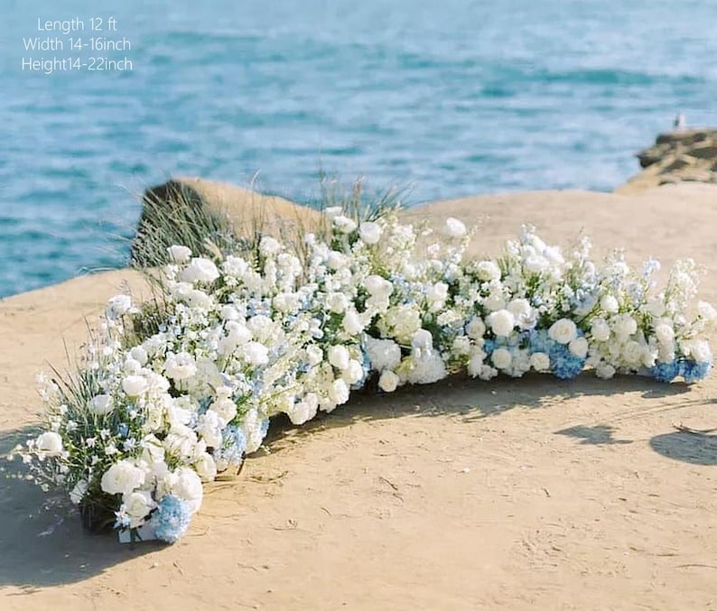 May include: A floral arrangement featuring white and blue flowers, with a length of 12 ft, a width of 14-16 inches, and a height of 14-22 inches. The flowers are arranged on a sandy surface with the ocean in the background.