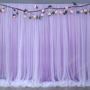 Pode incluir: Um fundo feito de tecido de tule branco com uma guirlanda de flores cor-de-rosa e folhas verdes drapeadas no topo. O tule é drapeado de uma forma que cria um efeito suave e fluido. O fundo é perfeito para um casamento, uma festa ou outro evento especial.