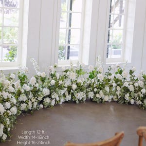 Op de afbeelding: Een witte bloemenboog met groen, met een lengte van 3,66 meter, een breedte van 35,56 tot 40,64 centimeter en een hoogte van 40,64 tot 61 centimeter. De boog is gemaakt van witte bloemen en groen en is perfect voor een bruiloft of andere speciale gelegenheid.