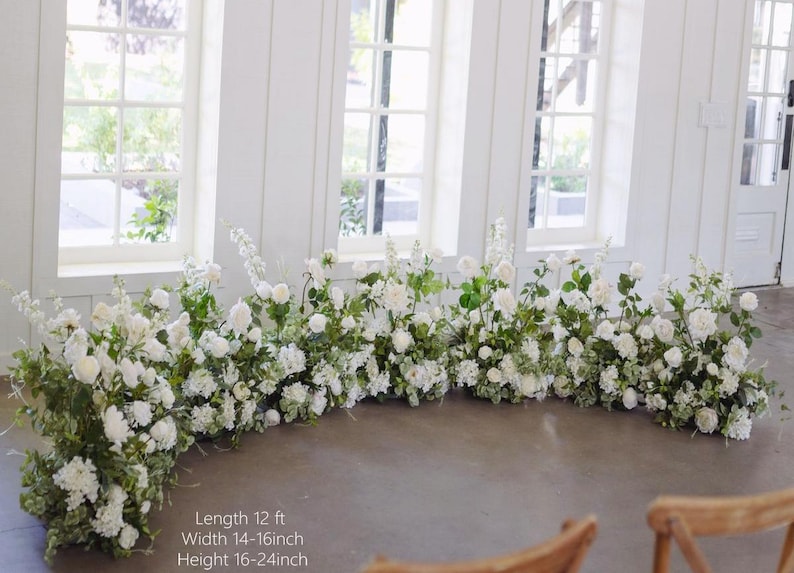 Op de afbeelding: Een witte bloemenboog met groen, met een lengte van 3,66 meter, een breedte van 35,56 tot 40,64 centimeter en een hoogte van 40,64 tot 61 centimeter. De boog is gemaakt van witte bloemen en groen en is perfect voor een bruiloft of andere speciale gelegenheid.