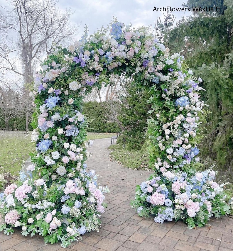 May include: A floral archway, adorned with an abundance of pastel-colored flowers, including white, pink, and blue blooms, creates a picturesque scene. The arch is lush with greenery and the text "ArchFlowers W8xH8 ft" is visible.
