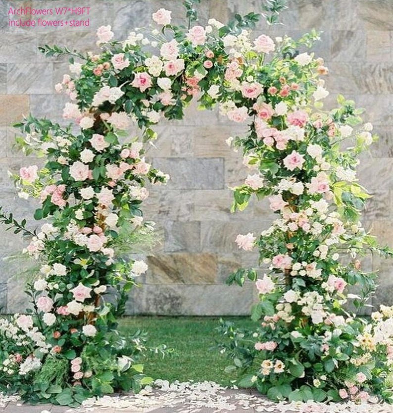 K&ouml;nnte beinhalten: Ein floraler Torbogen mit rosa und wei&szlig;en Rosen und gr&uuml;nem Laub. Der Bogen steht vor einer Steinmauer und gr&uuml;nem Gras. Der Text "ArchFlowers W7*H9FT include flowers + stand" ist sichtbar.