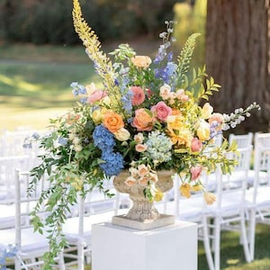 Op de afbeelding: Een groot bloemstuk in een decoratieve urn, ongeveer 51 cm breed. Het arrangement bevat een verscheidenheid aan kleurrijke bloemen, waaronder rozen, hortensia's en andere bloemen in tinten roze, oranje, geel en blauw. De urn staat op een witte sokkel.