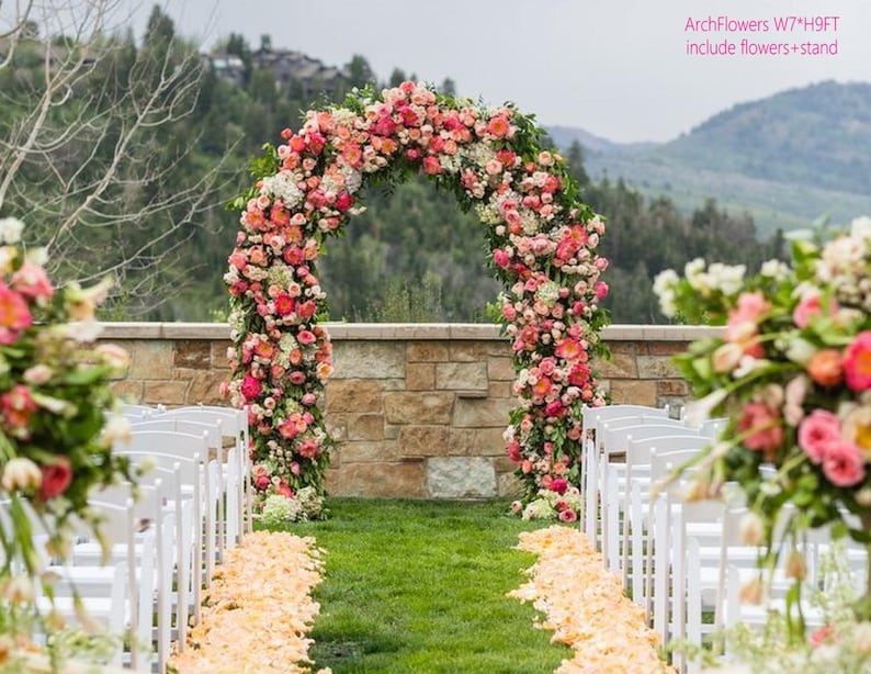 K&ouml;nnte beinhalten: Ein floraler Torbogen, etwa 213 cm breit und 274 cm hoch, geschm&uuml;ckt mit rosa, pfirsichfarbenen und wei&szlig;en Blumen, dient als Kulisse f&uuml;r eine Hochzeitszeremonie. Wei&szlig;e St&uuml;hle s&auml;umen den Gang, mit einem Bl&uuml;tenblattweg, der zum Torbogen f&uuml;hrt. Der Text "ArchFlowers W7*H9FT include flowers + stand" ist sichtbar.