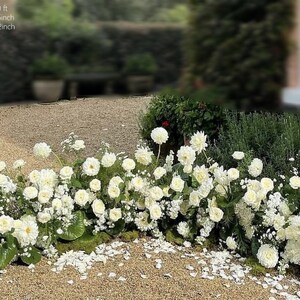 May include: A low, lush floral arrangement featuring white roses and other white flowers. The arrangement is approximately 10 feet long, 14-16 inches wide, and 14-22 inches high. The flowers are set against a backdrop of greenery and gravel.