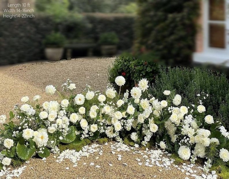 May include: A low, lush floral arrangement featuring white roses and other white flowers. The arrangement is approximately 10 feet long, 14-16 inches wide, and 14-22 inches high. The flowers are set against a backdrop of greenery and gravel.