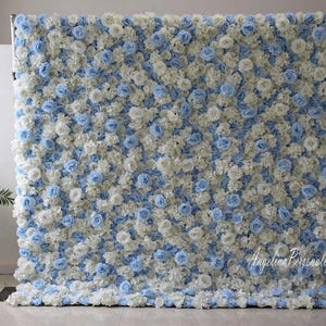 May include: A backdrop made of artificial flowers in white and light blue. The flowers are arranged in a dense pattern, creating a full and lush look. The backdrop is perfect for weddings, parties, or other special events.