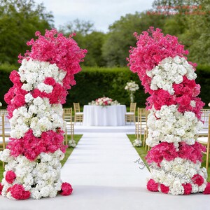 May include: Two floral arches, composed of white and hot pink flowers, frame a wedding aisle. The arches are made of a mix of white and pink flowers, with a dense arrangement. A white carpet leads to a table with a floral centerpiece.