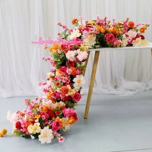 May include: A cascading floral arrangement of pink, orange, and yellow flowers, with greenery, draped over a white table with wooden legs. The flowers are artificial and create a beautiful and festive centrepiece.