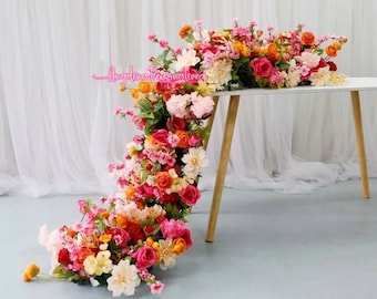 May include: A cascading floral arrangement of pink, orange, and yellow flowers, with greenery, draped over a white table with wooden legs. The flowers are artificial and create a beautiful and festive centerpiece.
