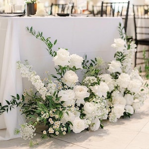 May include: A floral arrangement featuring white roses, delphiniums, and small daisies with green foliage. The flowers are clustered together in a cascading design, displaying various petal textures and shades of white, accented by thin green stems.