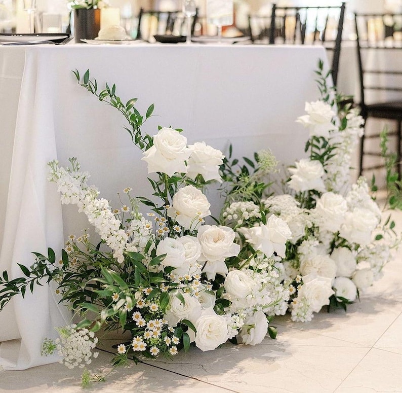 May include: A floral arrangement featuring white roses, delphiniums, and small daisies with green foliage. The flowers are clustered together in a cascading design, displaying various petal textures and shades of white, accented by thin green stems.