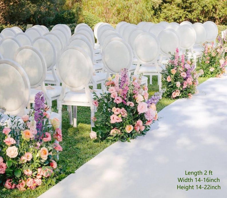 May include: Rows of white oval-back chairs arranged for an event, with floral arrangements of pink and purple flowers placed along a white aisle runner. The floral displays measure 2 feet in length, 14-16 inches in width, and 14-22 inches in height.