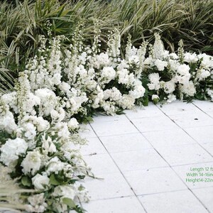 May include: A display of white flowers, including roses and other varieties, arranged on a white tiled surface. The arrangement is approximately 12 feet long, 14-16 inches wide, and 14-22 inches high. Green foliage provides a backdrop.
