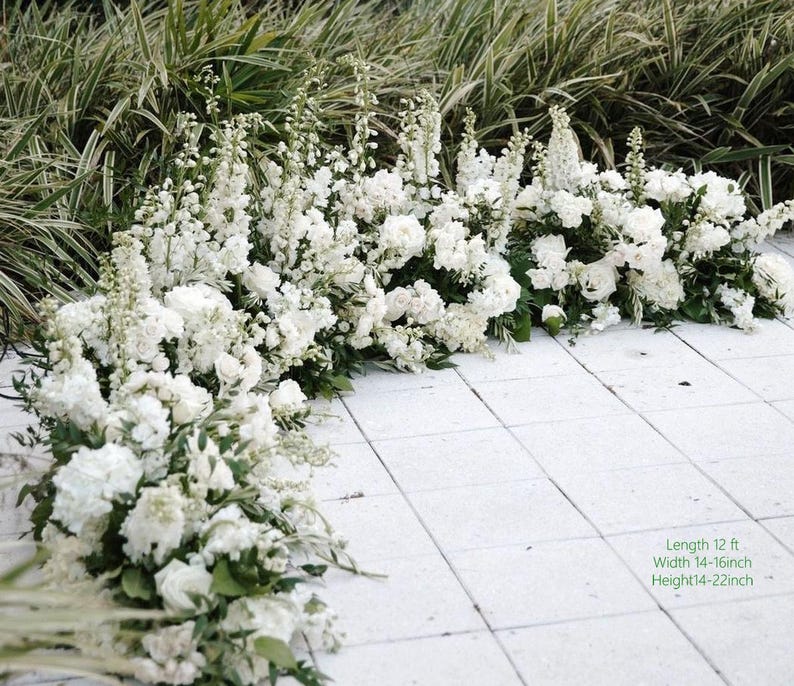May include: A display of white flowers, including roses and other varieties, arranged on a white tiled surface. The arrangement is approximately 12 feet long, 14-16 inches wide, and 14-22 inches high. Green foliage provides a backdrop.