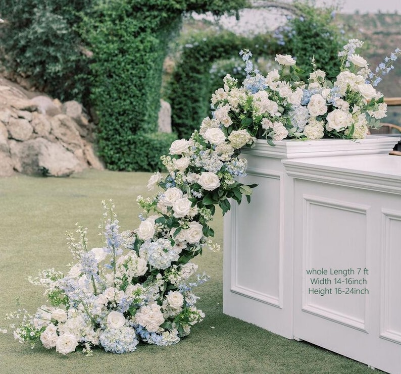 May include: A white structure decorated with a flowing floral display. The flowers are mainly white roses and blue hydrangeas, with green foliage. Text on the structure indicates a length of 2.13 m, width 35.5-40.6 cm, and height 40.6-61 cm.